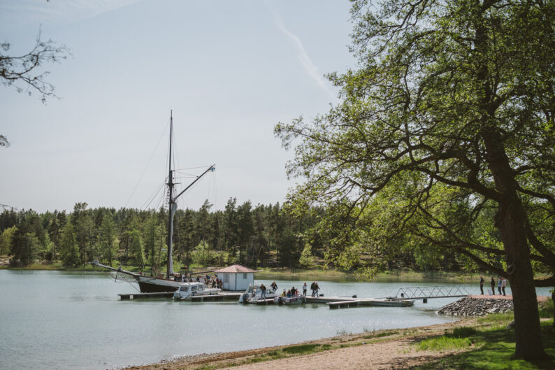 Historiallinen purjelaiva ja pienempiä veneitä laiturissa rauhallisessa lahdessa metsän ympäröimänä. Laiturilla liikkuu kävijöitä, ja suuri lehtipuu kehystää maisemaa. Aurinkoinen sää ja tyyni vesi luovat tunnelmallisen saaristomaiseman Söderlångvikin kartanossa.